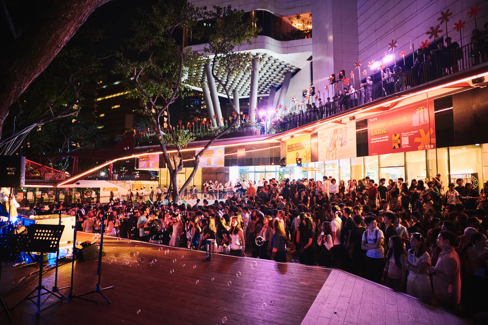 The finale: a crowd fills the *SCAPE plaza at night under purple light, with people lining every balcony above